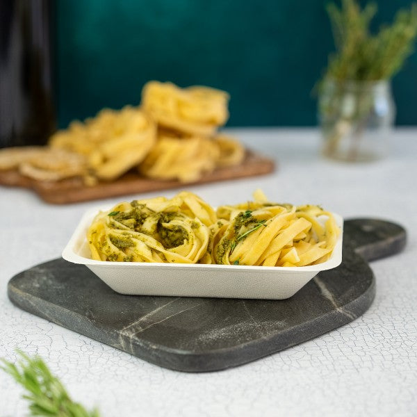 Pasta dish with green sauce in a white container on a dark stone board with a blurred background
