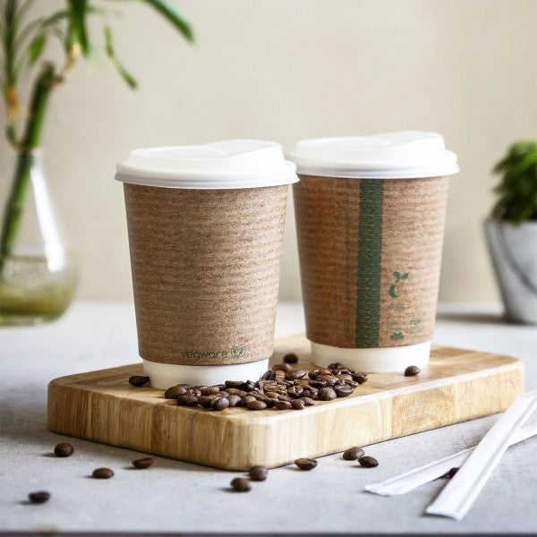Two disposable coffee cups on a wooden board with coffee beans scattered around, and a green plant in the background.